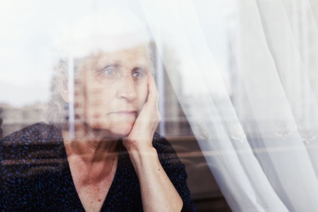 Image shows a pensive senior woman looking through a window and thinking