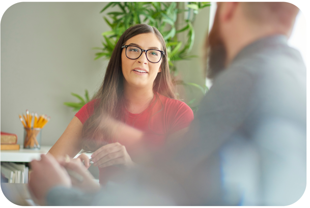 A woman in her 20s is sat down and looking towards a man whose back is to us and whose image is blurry. The woman is in a red t-shirt, has long brown hair and glasses. She looks supportive and kind and appears to be listening to the man and has a pen in her hand, as if taking notes. 