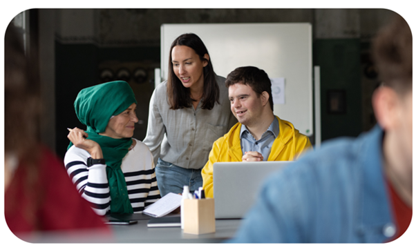 An image of 3 diverse people in what looks like a community setting. One of the individuals is standing slightly behind the other two who are sat down at a desk. The one standing is talking to them and looks kind and caring. There is one man in a bright yellow coat who has a laptop in front of him. The woman sat next to him has an emerald green head scarf. They are all smiling. 